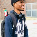Smiling young man wearing cap and headphones, standing with backpack outdoors.